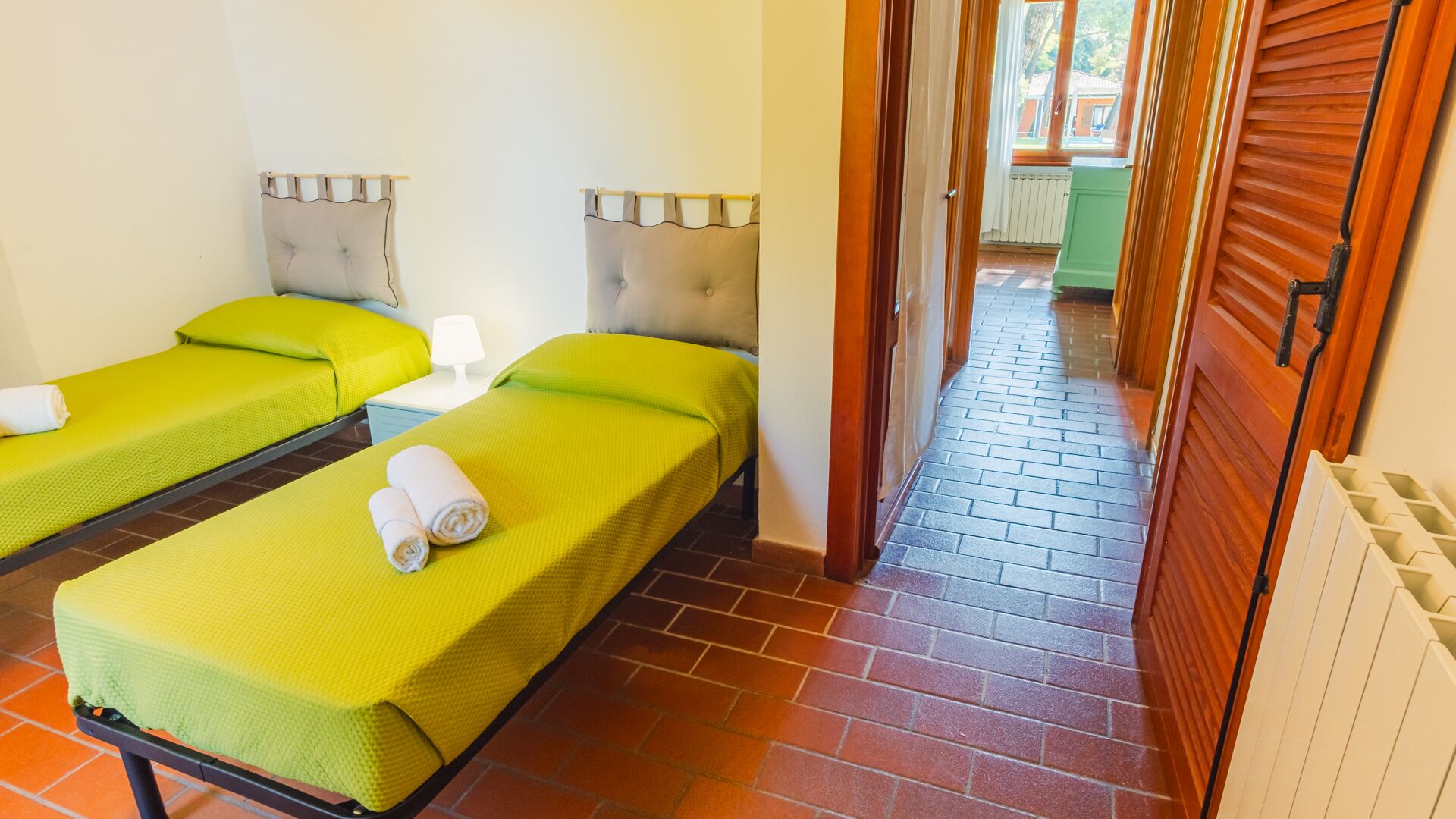 Ein Hotelzimmer mit zwei Einzelbetten, einem kleinen Tisch mit einer Lampe und Blick ins Freie durch ein Fenster.