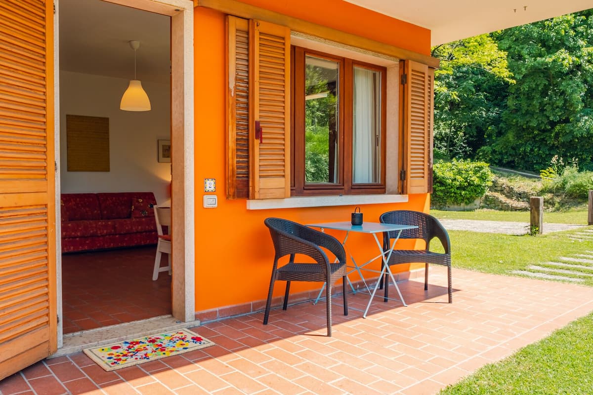 Une porte ouverte donne sur une pièce où se trouvent un canapé rouge, une petite table et deux chaises en osier, placées à l’extérieur.