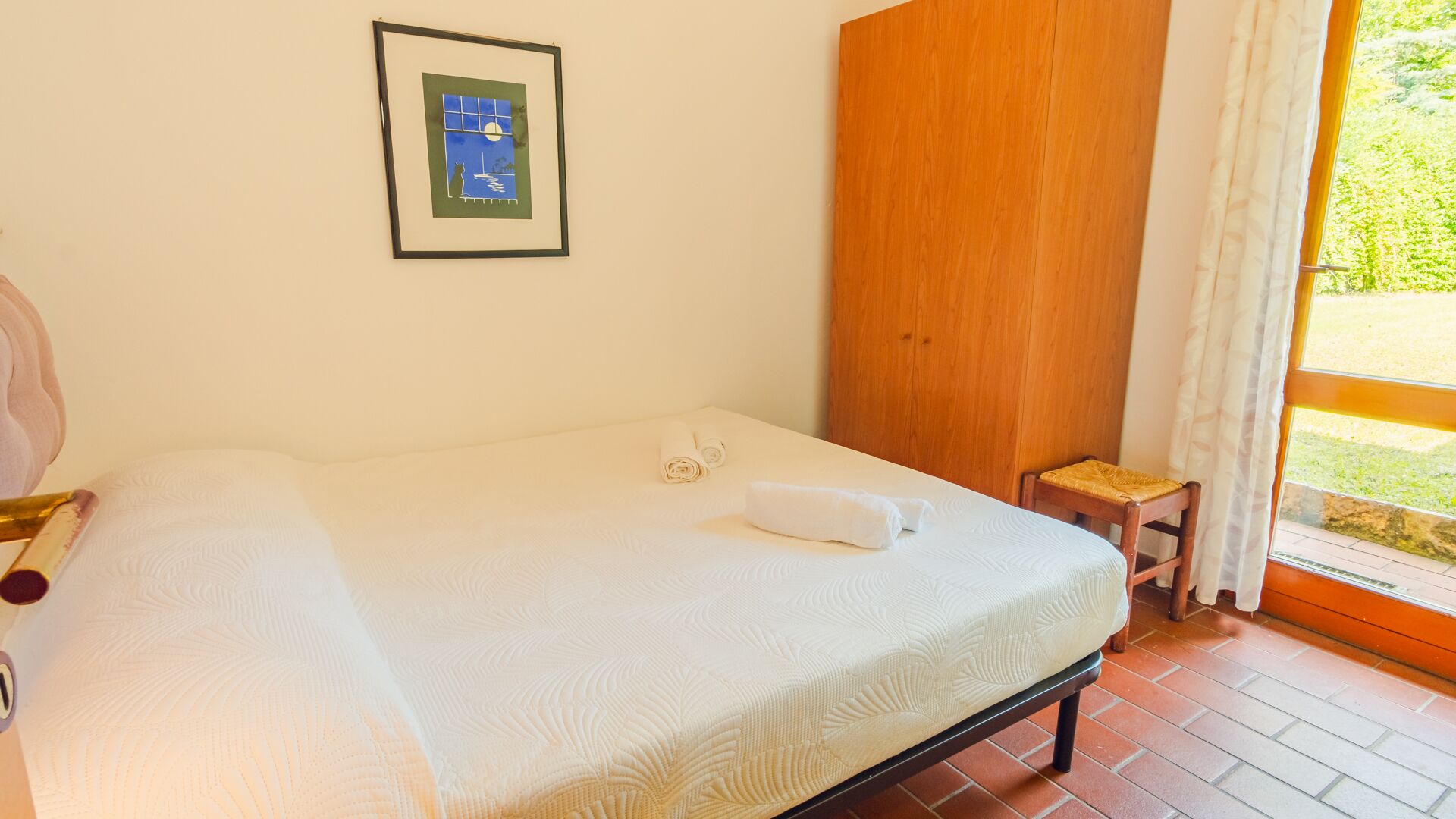 A neatly made bed with a white comforter and a rolled towel, a framed picture on the wall, and a wooden door.