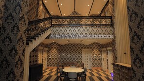 A room with a checkered floor, patterned wallpaper, a round table with chairs, and a staircase with ornate railings. - THE ROYAL VENETIAN (Samalkha)