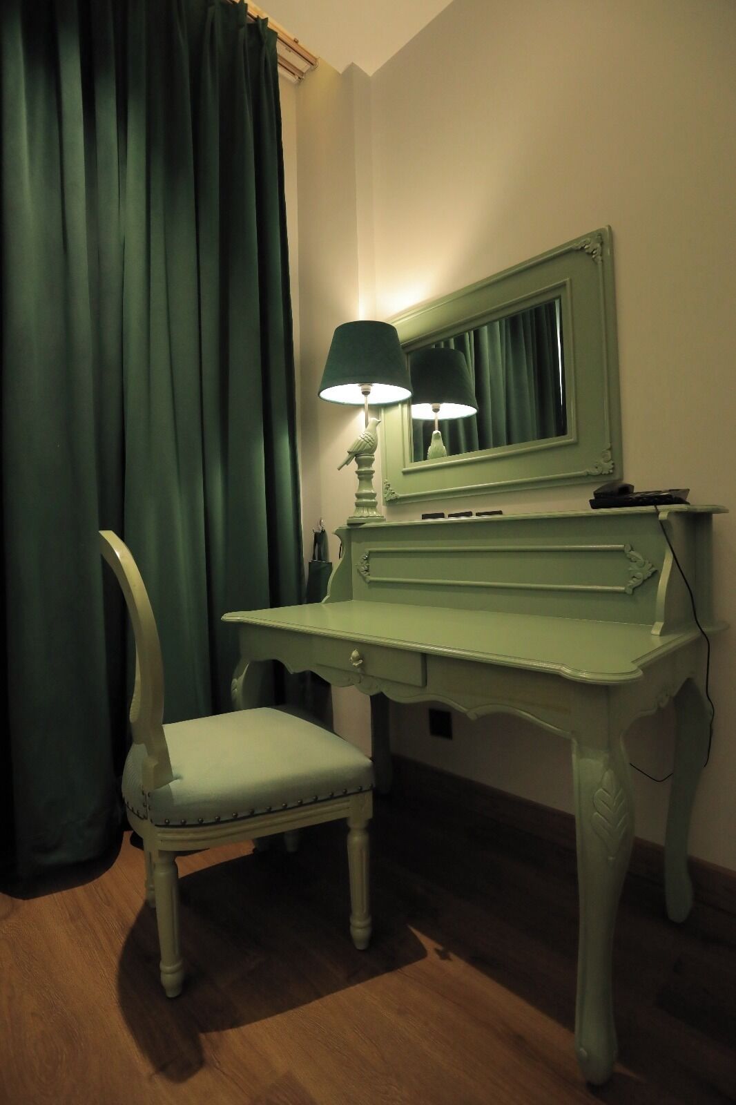 Eine grüne Klavierbank mit einer Lampe, einem Spiegel und einem Stuhl in einem Raum mit Holzboden und grünen Vorhängen.