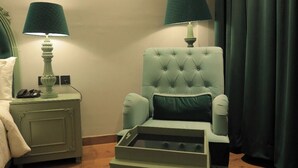 A room with a tufted armchair, a small table, two bedside tables with lamps, and a dark curtain. - THE ROYAL VENETIAN (Samalkha)