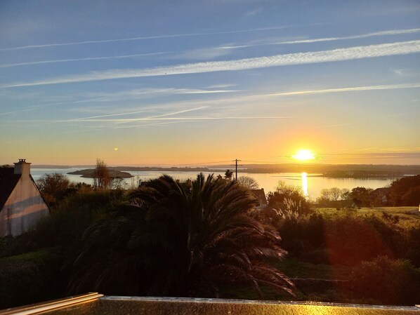 A sunset over a body of water with a distant shoreline, palm trees, and a building with a sloped roof.