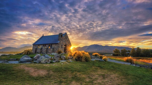 Ein Gebäude aus Stein mit einem geneigten Dach, umgeben von Gras und Steinen, mit Bergen im Hintergrund und einem Sonnenuntergang.