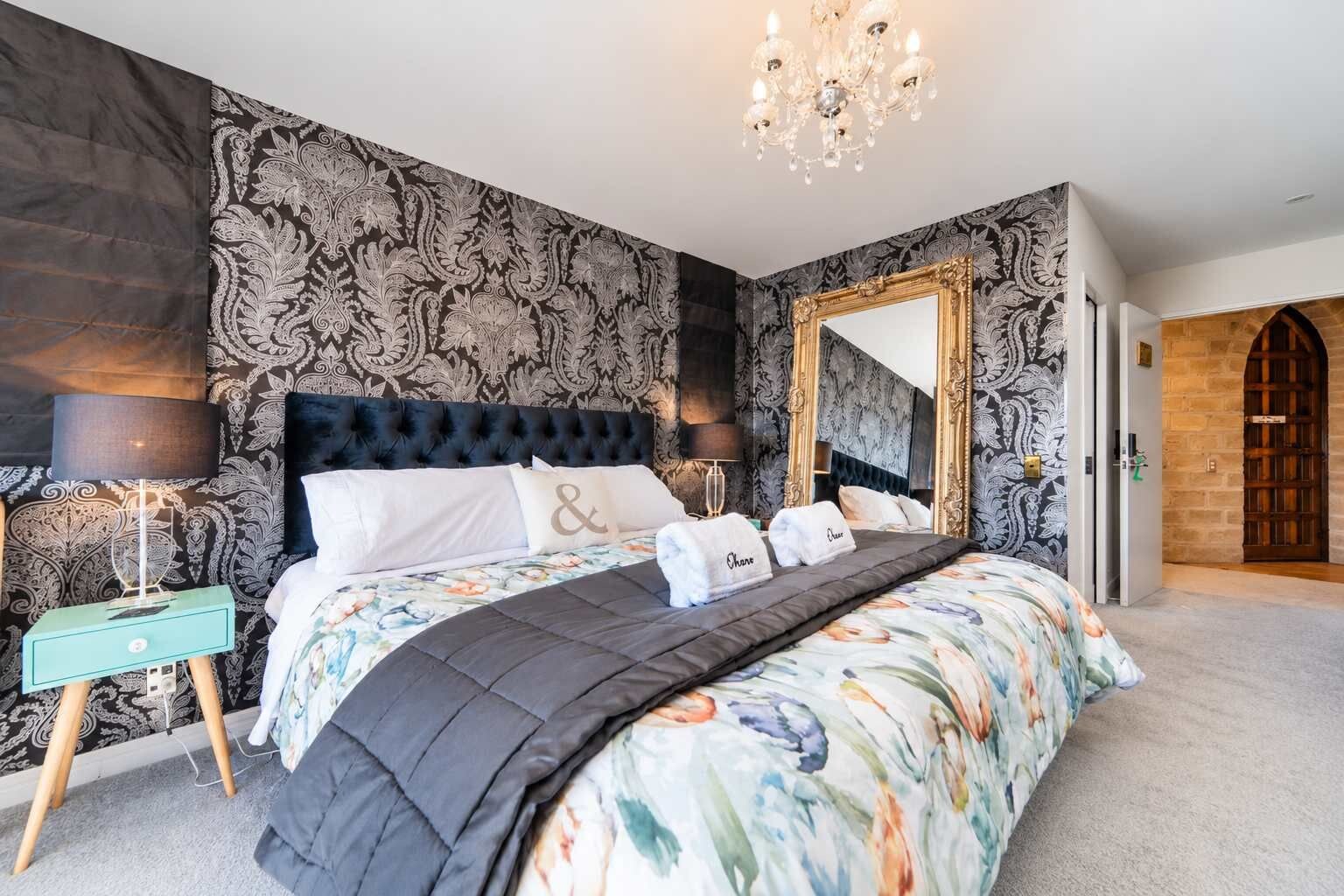 A bedroom with a large bed, two bedside tables, a chandelier, a mirror, and a patterned wallpaper.
