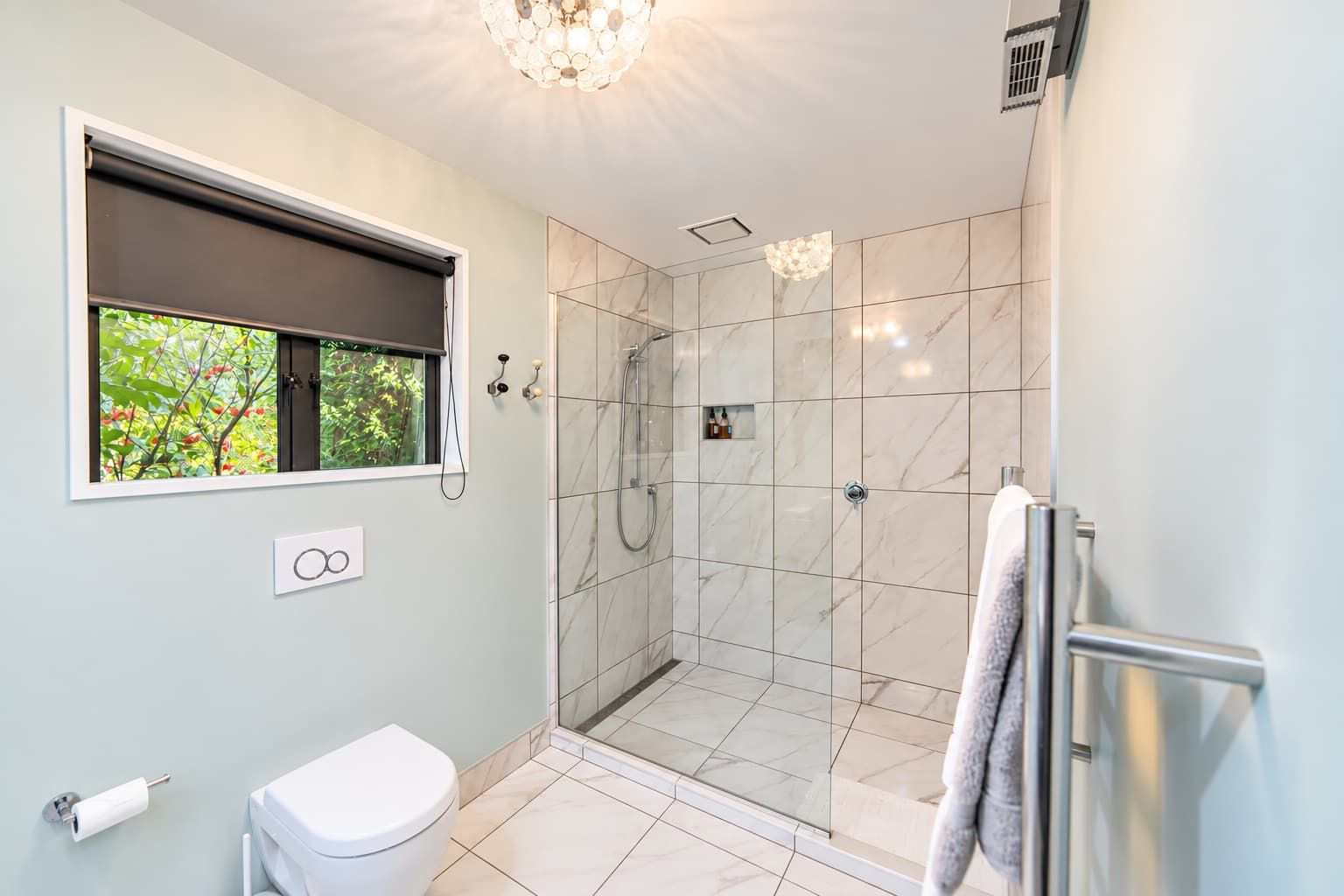 A modern bathroom with a shower, toilet, and a window with a view of greenery.