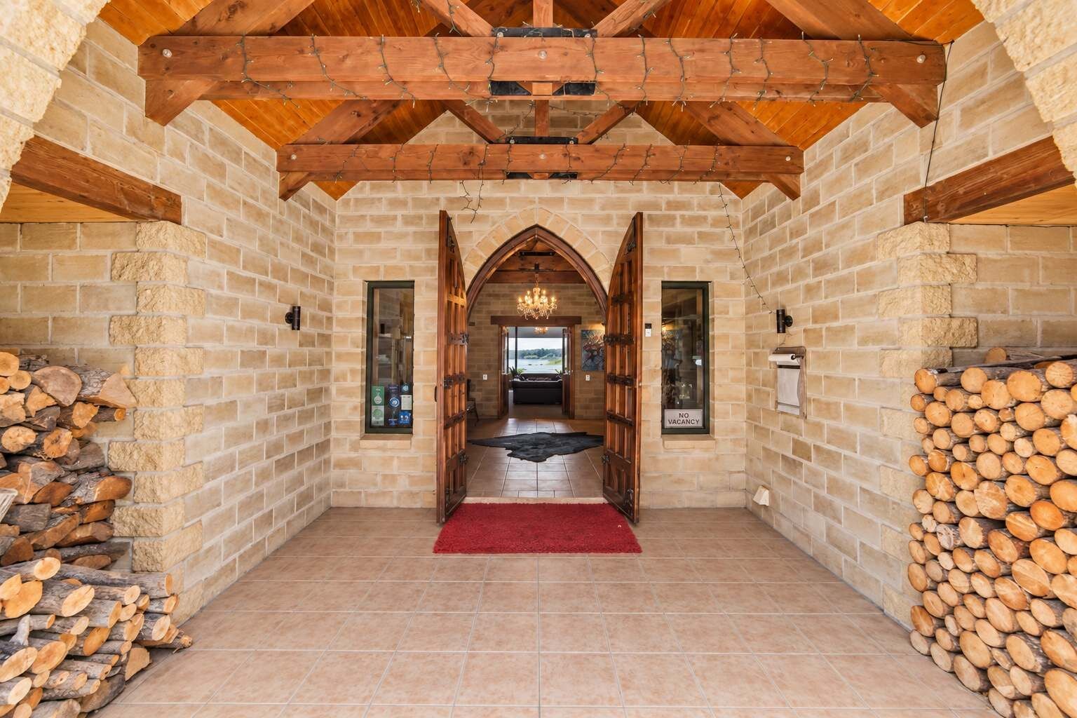 A spacious hallway with a wooden ceiling, stone walls, and a red carpet leading to a room with a chandelier.