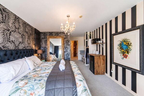A hotel room with a large bed, a chandelier, a mirror, a small kitchenette, and a framed artwork on the wall.