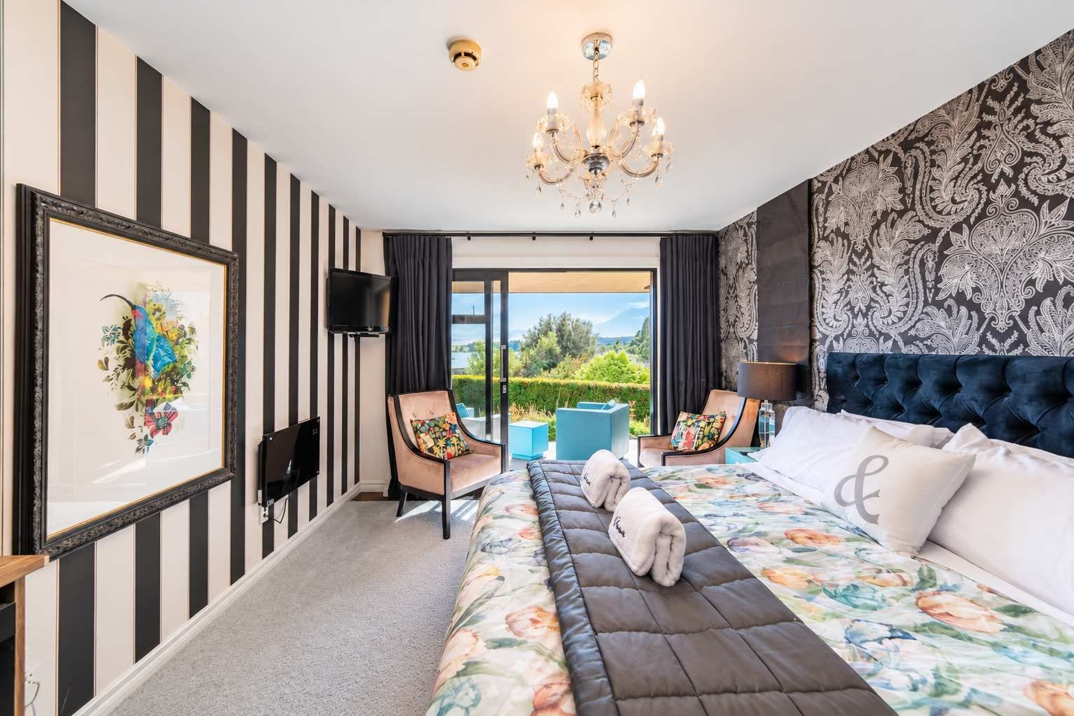 A hotel room with a large bed, a TV, a chandelier, a view of the outdoors, and a patterned wall.