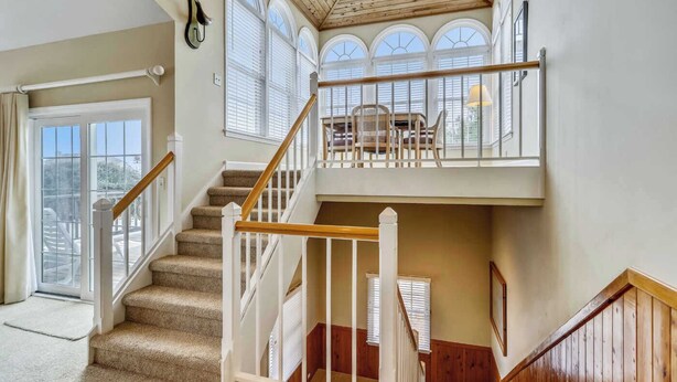 A multi-level interior with a carpeted staircase, wooden railings, and large windows with white shutters. - Cheven, Ships Watch Community in Duck, NC (Kitty Hawk)