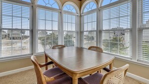 A dining area with a wooden table and four chairs, large arched windows with blinds, and a view of houses outside. - Cheven, Ships Watch Community in Duck, NC (Kitty Hawk)