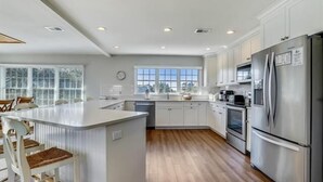 A modern kitchen with white cabinetry, a central island, stainless steel appliances, and a large window providing natural light. - Little, Ships Watch Community in Duck, NC (Kitty Hawk)