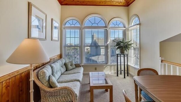 A cozy room with a wooden ceiling, a cushioned bench, a table, and a view through large arched windows. - Little, Ships Watch Community in Duck, NC (Kitty Hawk)