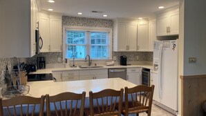 A modern kitchen with white cabinets, a large island with chairs, a window with blinds, and a refrigerator. - Shabedge, Ships Watch Community in Duck, NC (Kitty Hawk)