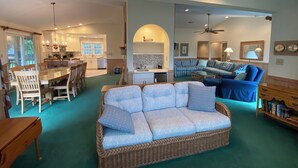A spacious living area with a large wicker sofa, a dining table with chairs, a kitchen in the background, and a ceiling fan. - Shabedge, Ships Watch Community in Duck, NC (Kitty Hawk)
