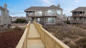 A wooden boardwalk leading to a beachfront property with multiple-story buildings, balconies, and a clear sky. - Spendlove, Ships Watch Community in Duck, NC (Duck)