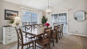 A dining area with a large wooden table, six chairs, a white cabinet, a potted plant, and a hanging light fixture. - Spendlove, Ships Watch Community in Duck, NC (Duck)