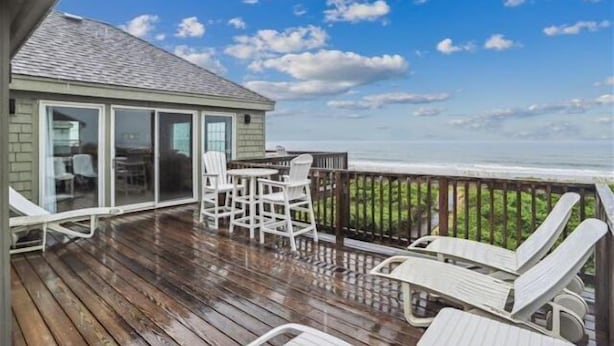 A wooden deck with white chairs and a table, a glass door leading to a room, and a view of the ocean. - Spendlove, Ships Watch Community in Duck, NC (Duck)