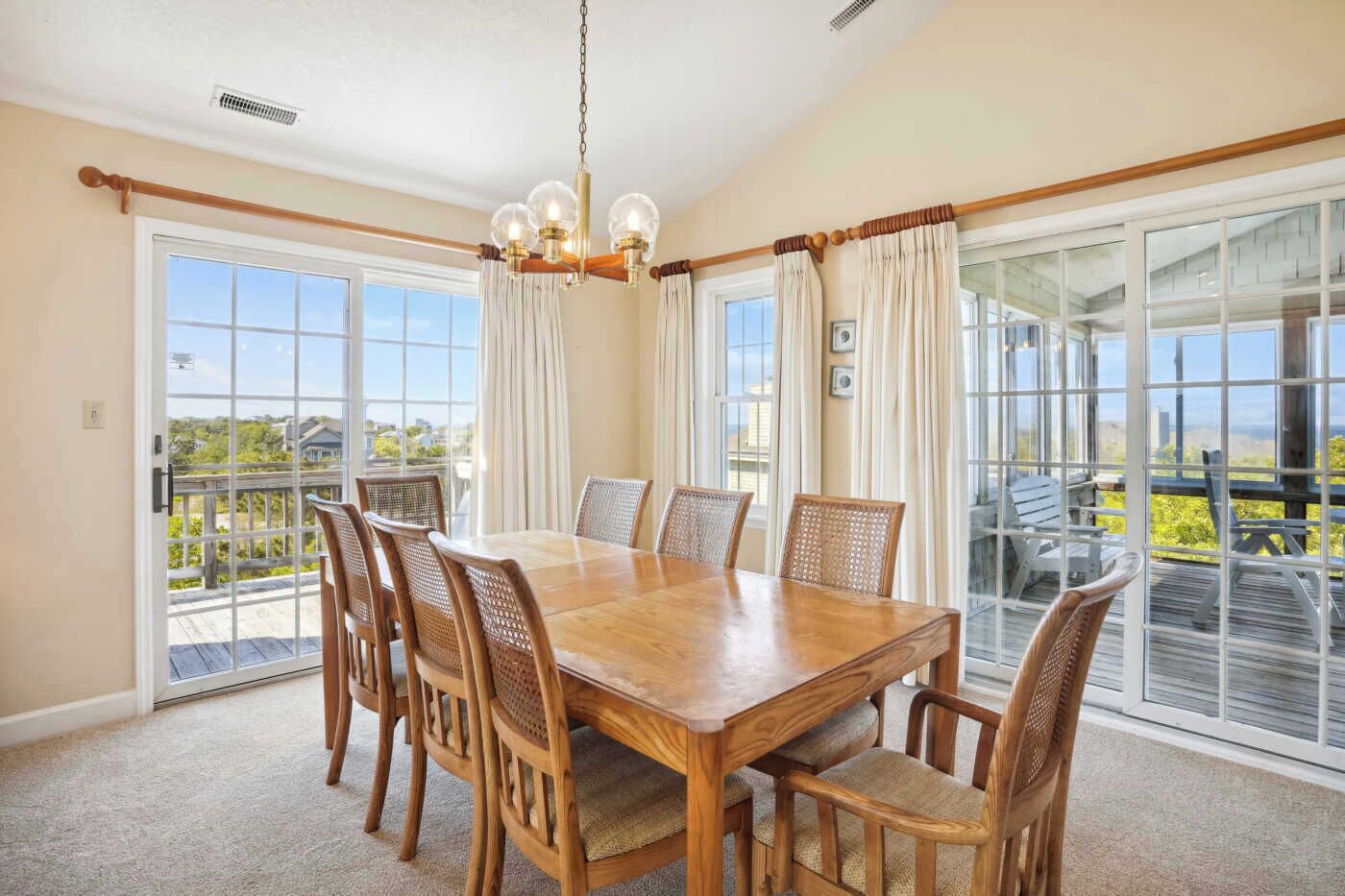 Une salle à manger avec une table et des chaises en bois, de grandes fenêtres avec des rideaux et une vue sur l’extérieur.