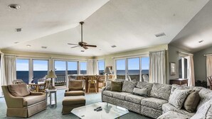 A spacious living room with a large sofa, armchairs, and a glass coffee table. There is a ceiling fan, large windows with ocean views, and a balcony with outdoor seating. - Hemmington, Ships Watch Community in Duck, NC (Duck)