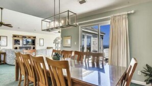 A dining area with a glass-top table and wooden chairs, a ceiling fan, and a view of the ocean through a sliding glass door. - Hemmington, Ships Watch Community in Duck, NC (Duck)