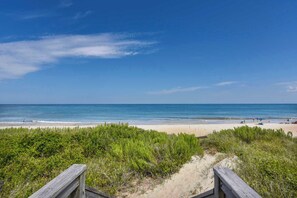 Una playa con un paseo marítimo de madera que conduce a la orilla, cielo azul despejado y mar tranquilo.