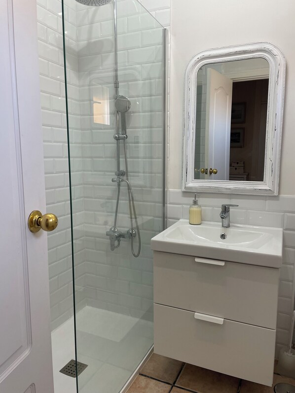 Kylpyhuoneessa on lasiseinällä varustettu suihku, valkoinen pesuallas, jonka yläpuolella on peili, sekä kultaisella kahvalla varustettu ovi.