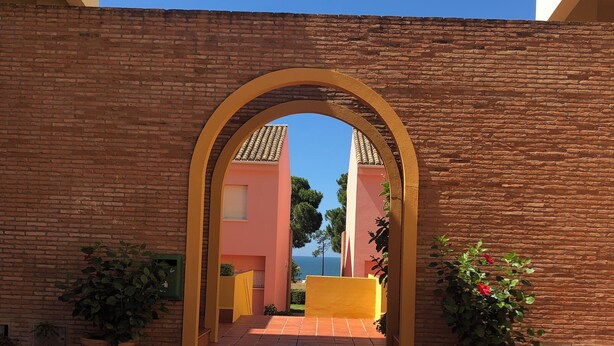 Un arco de ladrillo que conduce a un patio empedrado con plantas en macetas y una vista despejada de un edificio al fondo.