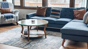Ein Wohnzimmer mit einem blauen Sofa, einem Glastisch und Blick auf eine Terrasse durch große Fenster.