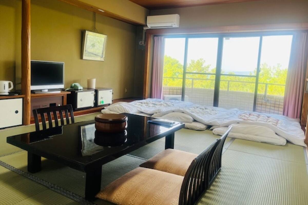Ett hotellrum med en säng, ett skrivbord med en dator, en stol, ett bord med en tekanna och ett fönster med utsikt över grönområden.