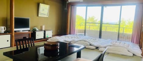 Et hotelværelse med seng, skrivebord med computer, stol, bord med te og et vindue med udsigt til grønne omgivelser.