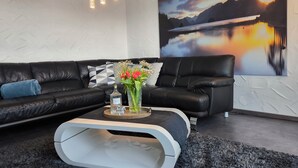 Un soggiorno moderno con un divano in pelle nera, un tavolino rotondo con un vaso di fiori e un grande dipinto paesaggistico appeso al muro.