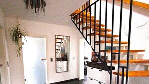 Un'ampia zona giorno con pavimento scuro, una scala con gradini in legno e ringhiere nere, una porta bianca e una pianta pensile.