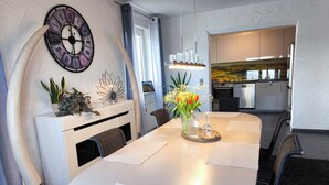 Un'area pranzo moderna con un tavolo bianco, sedie nere, un orologio decorativo e una cucina sullo sfondo.