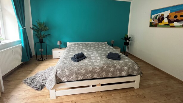 A bedroom with a teal wall, a bed with a patterned bedspread, two bedside tables with lamps, a potted plant, and a painting on the wall.