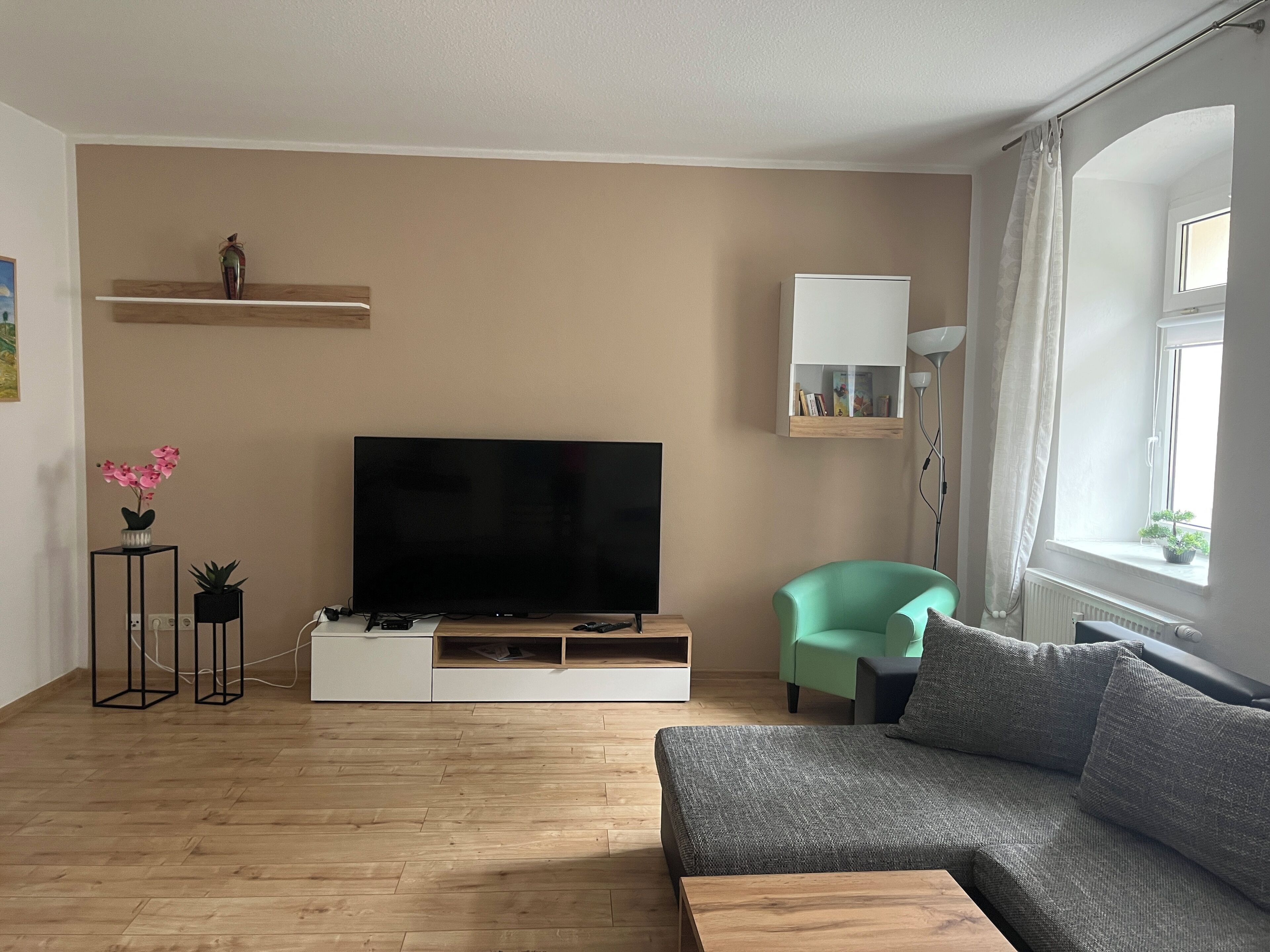 A modern living room with a flat-screen TV, a teal armchair, a grey sofa, and a wooden coffee table.