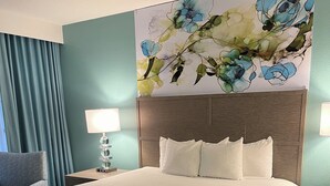 Habitación de hotel con una cama grande, cabecera con un mural floral, un sillón gris y una mesita de noche con lámpara.