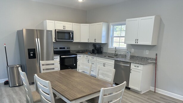 A modern kitchen with white cabinetry, a stainless steel refrigerator, a microwave, and a dining area with a wooden table and white chairs. - 2BR The Bandit Suite (Lexington)