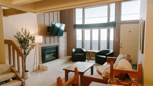 A living room with a fireplace, a TV mounted on the wall, a dining area with a table set for four, and a staircase leading to another level. - Budget Ski-In/Out+Walk to Dining (Steamboat Springs)
