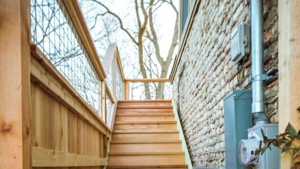 Una escalera de madera que sube hacia una ventana, con un muro de ladrillo a un lado y una valla de madera al otro.