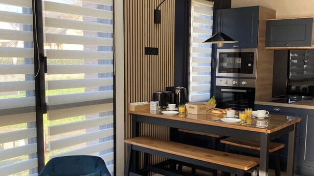 Une cuisine moderne avec un coin repas, comprenant une table en bois, deux chaises bleues et une grande fenêtre avec des stores.