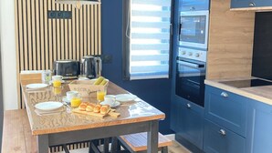 Une cuisine moderne avec une table en bois mise pour le petit-déjeuner, un four encastré et une fenêtre équipée de stores.
