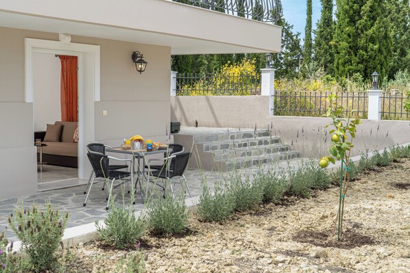 Un espace extérieur aménagé en terrasse, avec une table et des chaises, un petit jardin avec des plantes et une vue sur une clôture et des arbres.