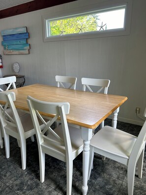 Comedor con mesa de madera y sillas blancas, una ventana decorada con motivos de estrellas de mar y un plato colgado en la pared.