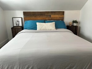 Un dormitorio con cama, cabecera de madera, mesita de noche con planta y un cuadro en la pared.
