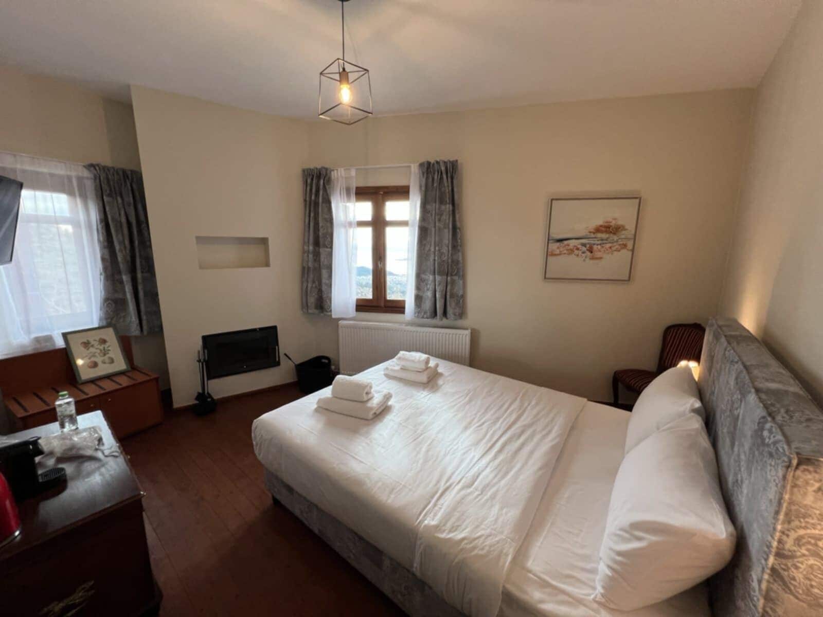 Ein Hotelzimmer mit einem Bett, einem Schreibtisch, einem Sessel, einem Fernseher, einem Fenster mit Vorhängen und einem Bild an der Wand.