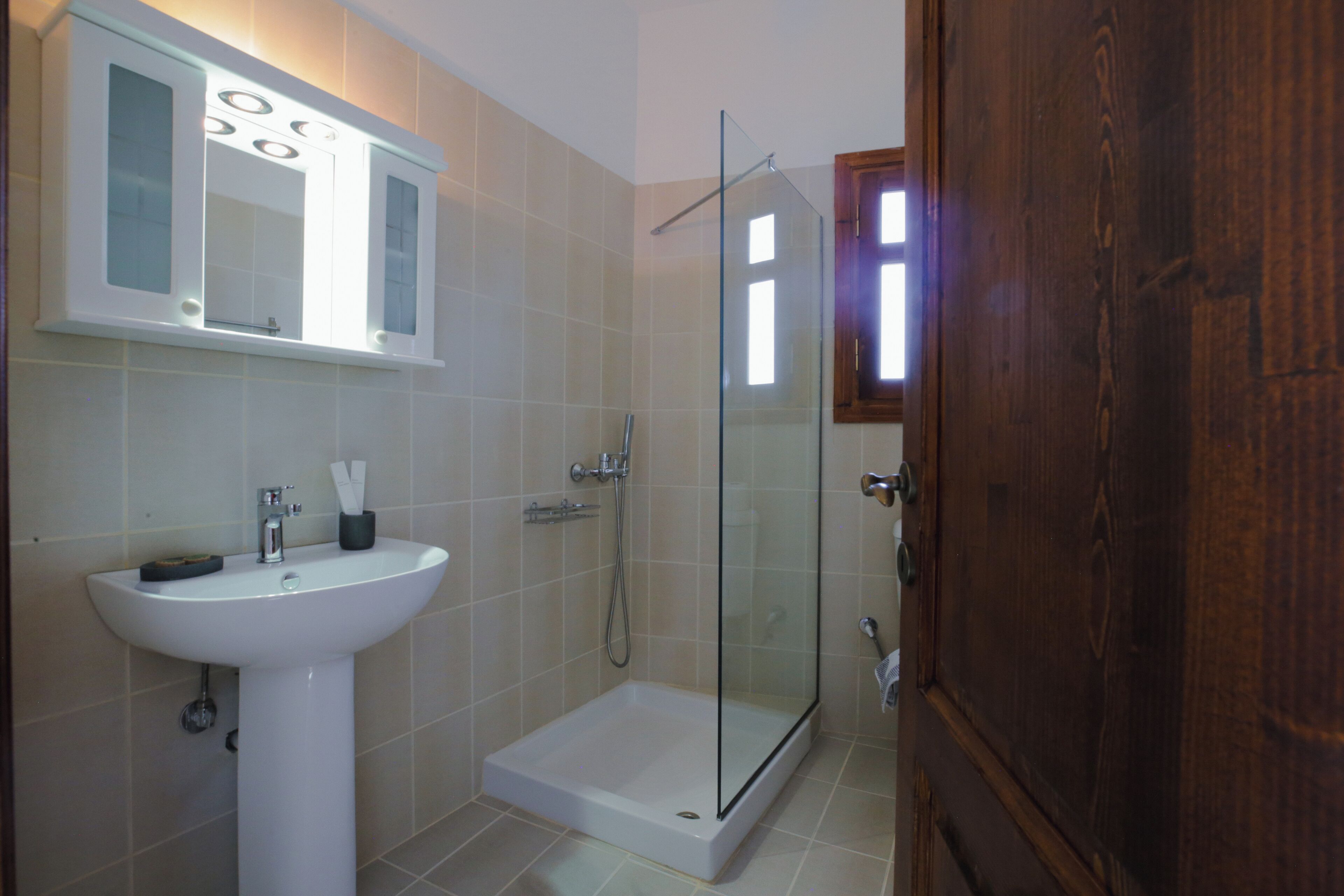 Un baño con un lavabo blanco, ducha con mampara de vidrio y puerta de madera.