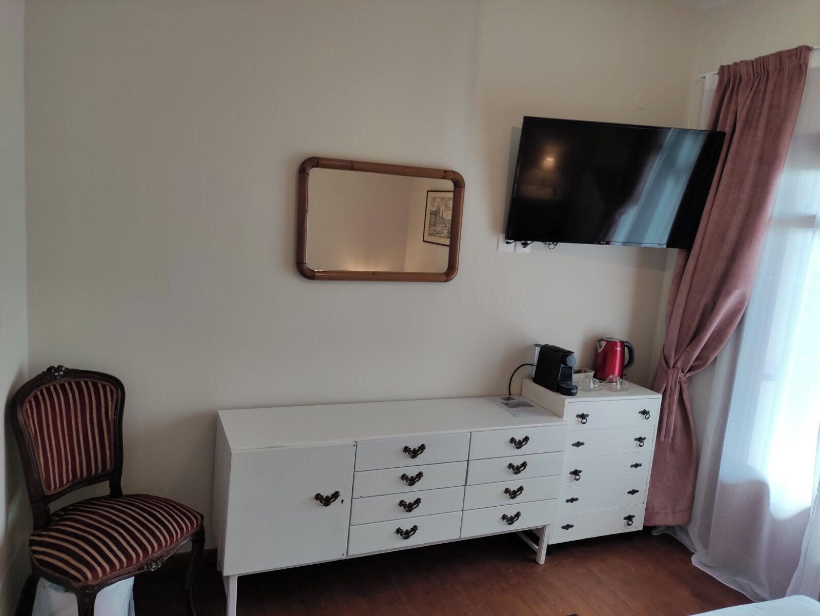 Une pièce comprenant une commode blanche à plusieurs tiroirs, une chaise avec un coussin à rayures, un téléviseur à écran plat fixé au mur et une fenêtre avec des rideaux.