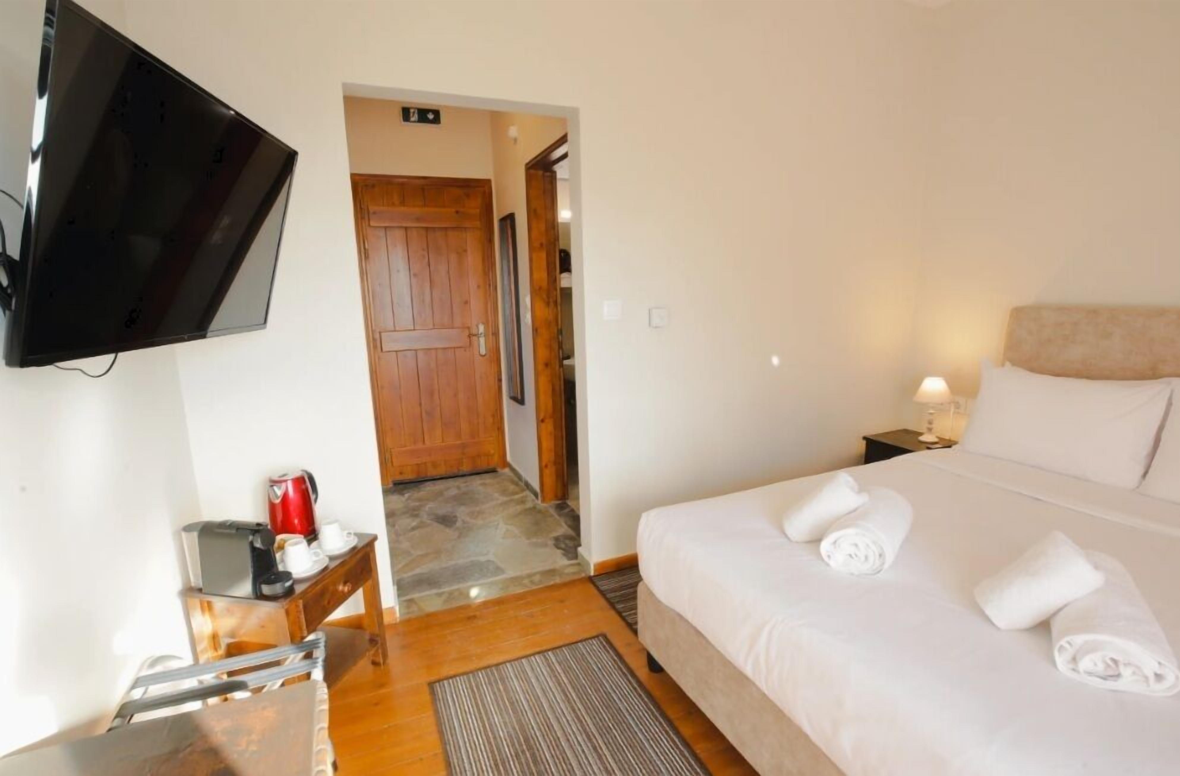 A hotel room with a bed, a TV mounted on the wall, a wooden door, a nightstand with a lamp, and a small table with a coffee maker.