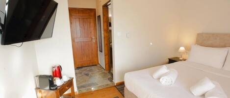 A hotel room with a bed, a TV mounted on the wall, a wooden door, a nightstand with a lamp, and a small table with a coffee maker.